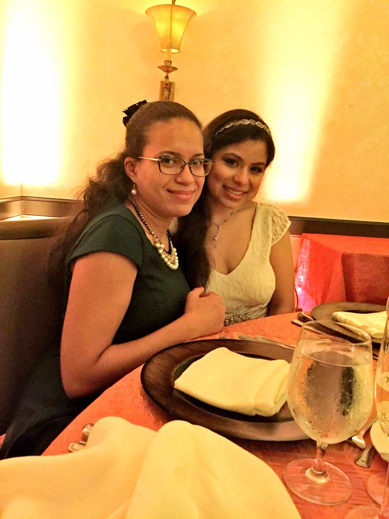 Two brunettes, one in a wedding gown, sit close together smiling.