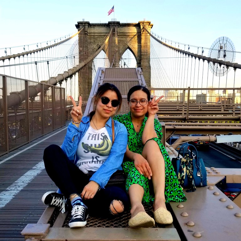 Two brunettes flash peace signs sitting on the Brooklyn Bridge.