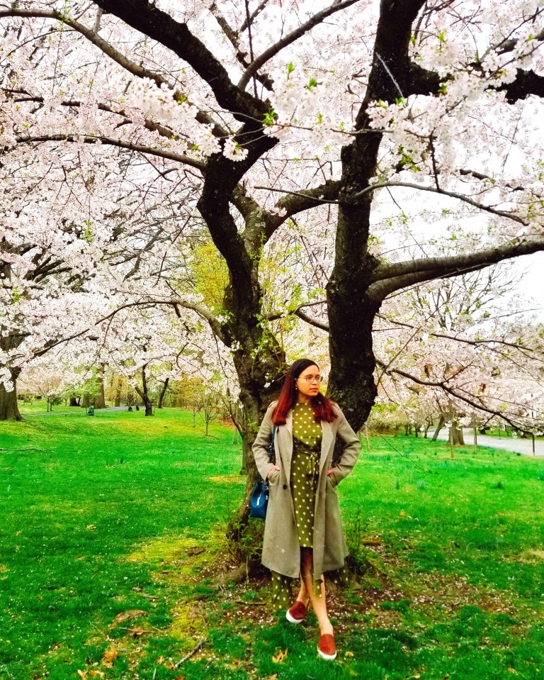 Brunette tucks hands in trench coat pockets in front of cherry blossom tree.