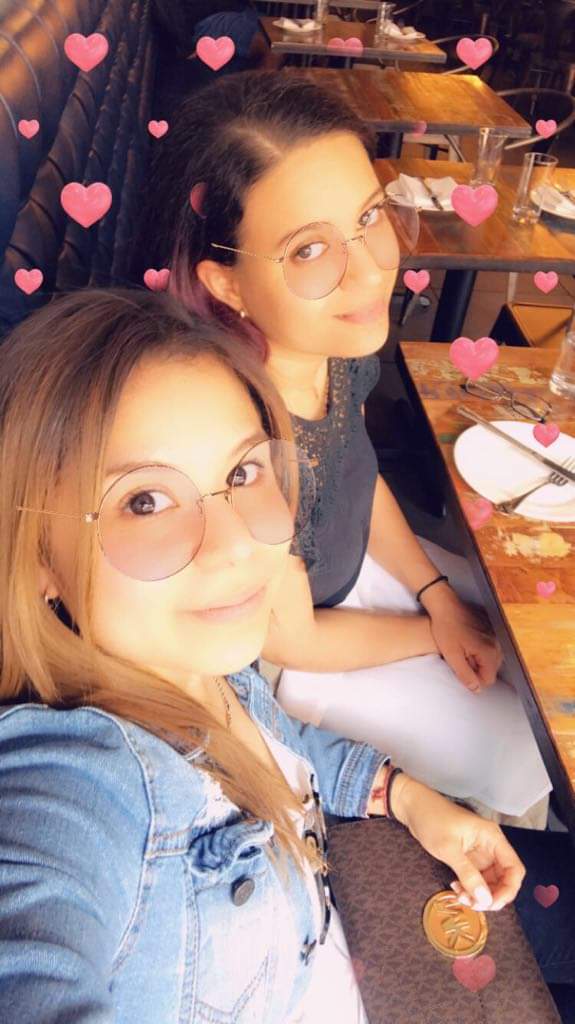 Two brunettes smile, round glasses and hearts filter overlaid on photo.