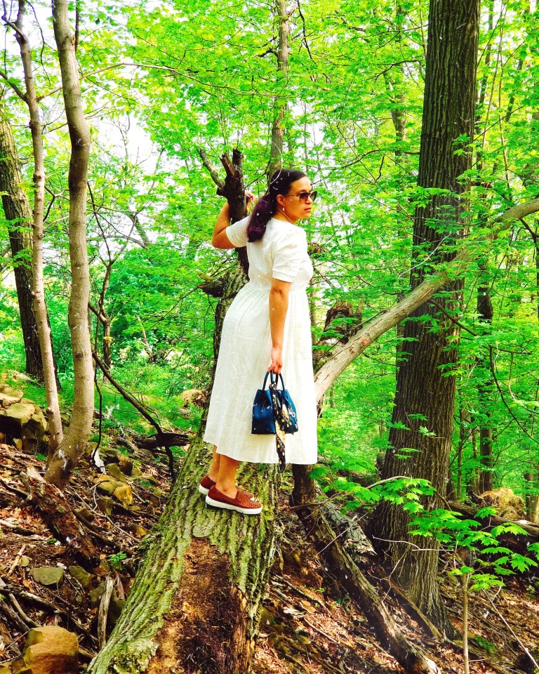 Brunette in white midi dress and cognac leather sneakers leans forward on fallen tree trunk, steadying herself with one hand on branch.