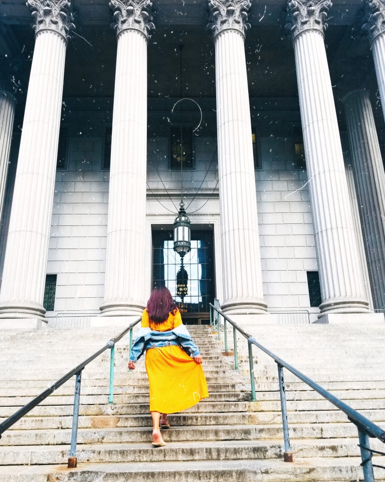 Brunette in yellow midi dress and denim jacket climbs steps of New York County Supreme Court, tall white columns and blocks, a tiled glass door and black lantern suspended from black chains.