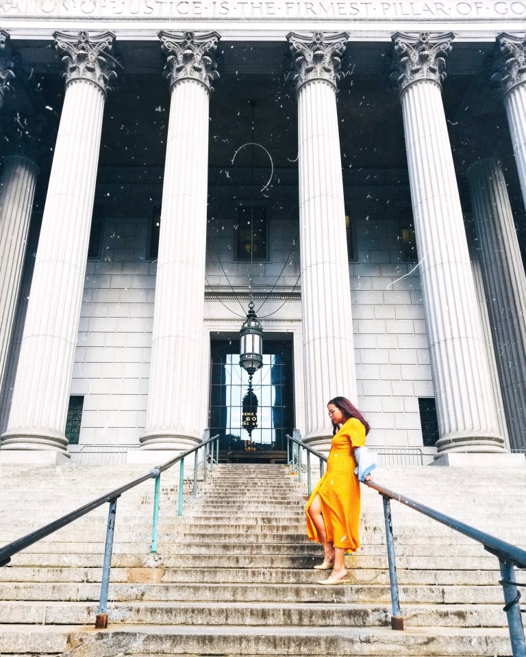 Brunette in yellow midi dress and denim jacket leans against railing at New York County Supreme Court, tall white columns and blocks, a tiled glass door and black lantern suspended from black chains.