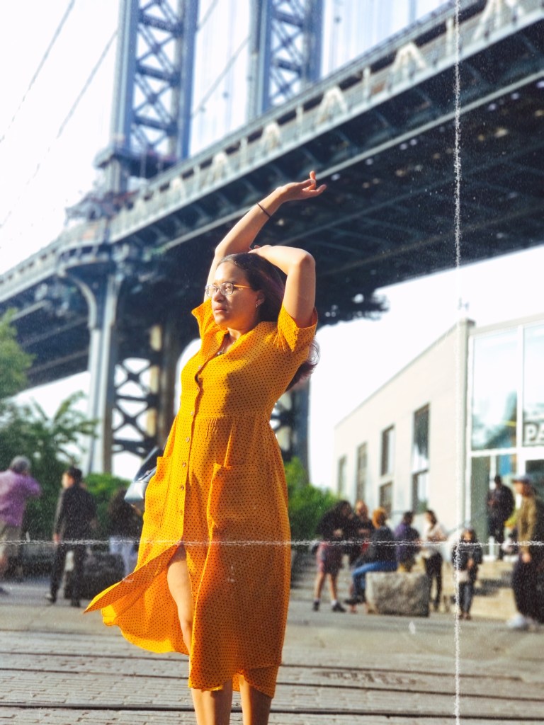 Brunette poses, one arm resting over her head, the other crooked up in the air, the Manhattan Bridge blurred in the background.