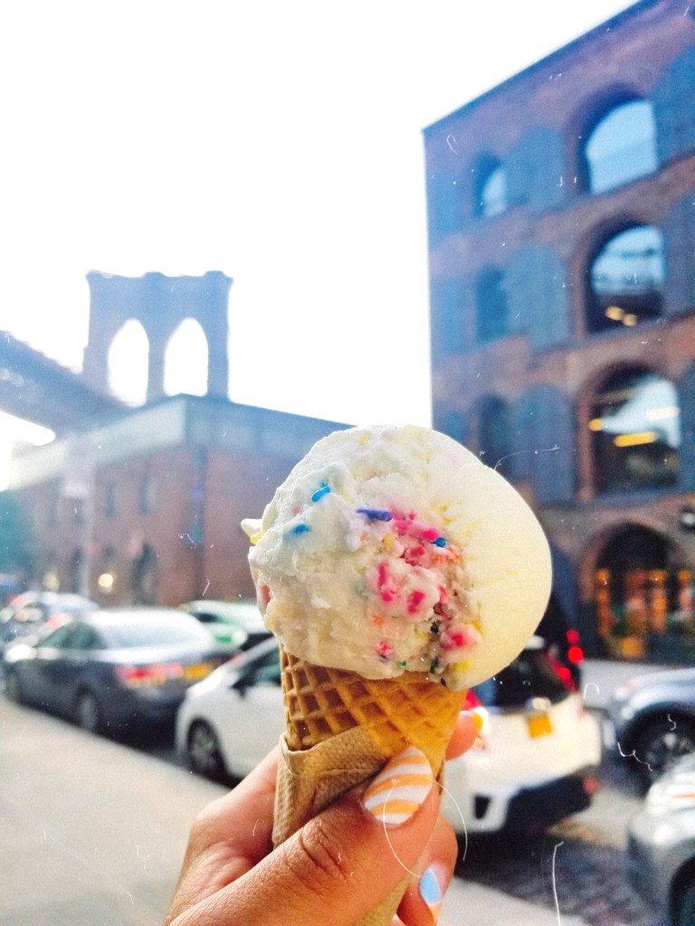 A manicured hand toasts the Brooklyn Bridge with a colorful scoop of ice cream.