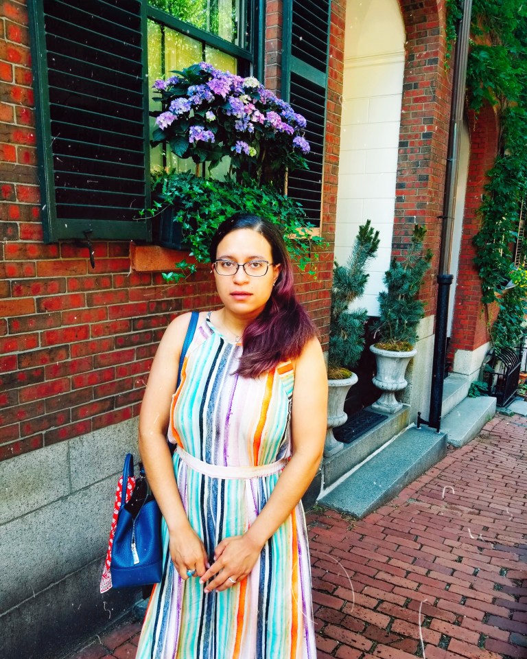 Brunette poses in colorful striped jumpsuit with brick row house, window box of blue and purple hydrangeas suspended over her head.