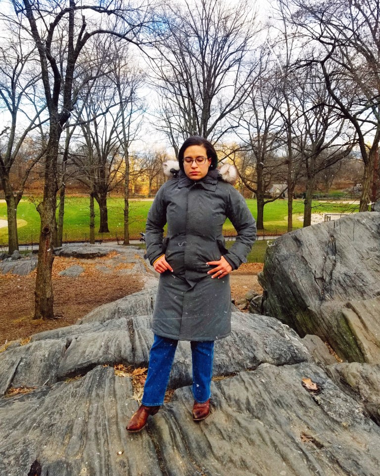 Brunette in a zipped-up gray parka stands on rock formation overlooking baseball field in Central Park.