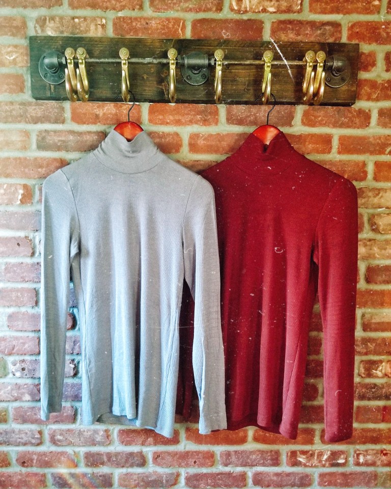 Two fitted turtleneck sweaters hang from rustic hooks against brick backdrop.