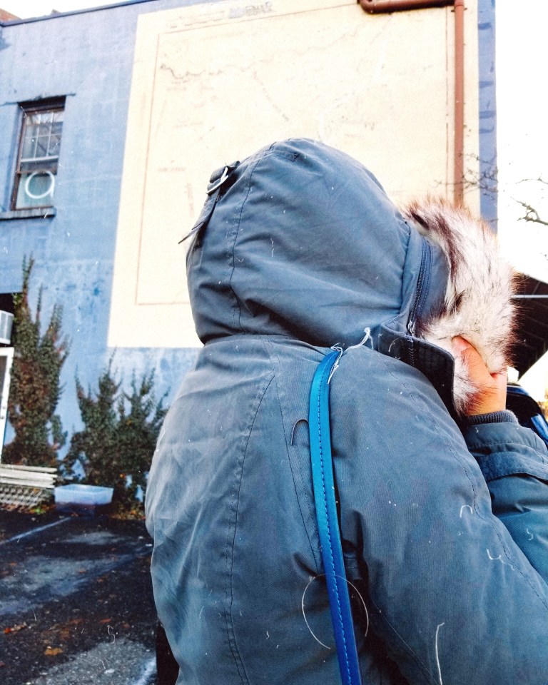 Woman stands with back turned to camera, gray furry hood pulled over her head.